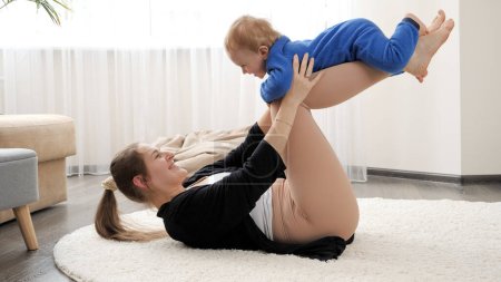 Photo for Happy smiling mother holding and rocking her baby son during fitness and yoga lesson at home. Family healthcare, active lifestyle, parenting and child development. - Royalty Free Image