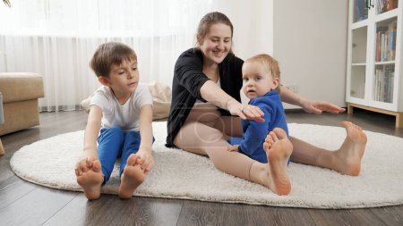 Téléchargez les photos : Jeune mère avec son bébé et son fils aîné faisant des exercices d'étirement et de fitness sur le tapis à la maison. Santé familiale, mode de vie actif, rôle parental et développement de l'enfant. - en image libre de droit