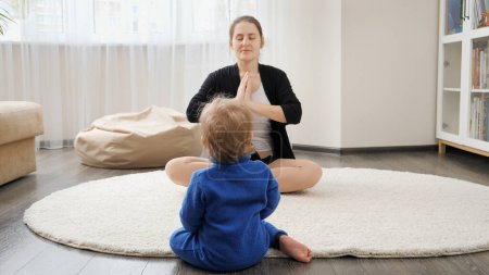 Foto de Pequeño niño mirando a su madre sentada en la alfombra y practicando yoga. Asistencia sanitaria familiar, estilo de vida activo, crianza y desarrollo infantil. - Imagen libre de derechos