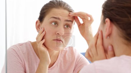 Photo for Brunette woman looking at her reflection in mirror and finding wrinkles on forehead. - Royalty Free Image