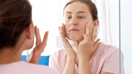 Photo for Happy smiling woman looking at her reflection in bathroom and checking her clear soft skin. Concept of female aging, skin issues, facial treatment and care - Royalty Free Image
