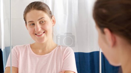 Téléchargez les photos : Portrait de femme brune souriante regardant son reflet dans le miroir de la salle de bain. - en image libre de droit