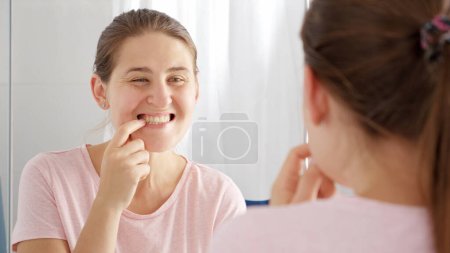 Téléchargez les photos : Portrait d'une jeune femme souriante à la recherche d'une plaque et de caries sur ses dents à la salle de bain. Concept de santé dentaire, auto-contrôle de la bouche et hygiène buccale - en image libre de droit