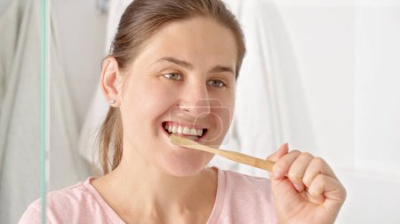 Foto de Mujer morena sonriente cepillándose los dientes con cepillo de dientes de madera en el baño. Concepto de salud dental, autocontrol bucal e higiene bucal - Imagen libre de derechos