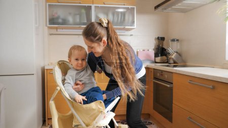 Photo for Young caring mother seating her baby in highchair on kitchen before giving breakfast. Concept of parenting, healthy nutrition and baby care. - Royalty Free Image