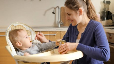 Foto de Portrait of smiling young mother feeding her baby son with fruit porridge. Concept of parenting, healthy nutrition and baby care. - Imagen libre de derechos