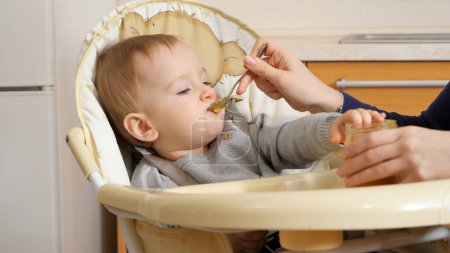 Foto de Closeup of little baby boy getting messy while eating porridge in highchair. Concept of parenting, healthy nutrition and baby care - Imagen libre de derechos