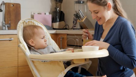 Foto de Retrato de madre sonriente con hijo pequeño sentado en la cocina en silla alta y esperando el desayuno. - Imagen libre de derechos