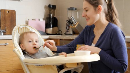 Téléchargez les photos : Portrait of smiling mother feeding her baby son with porridge from spoon. Concept of parenting, healthy nutrition and baby care - en image libre de droit