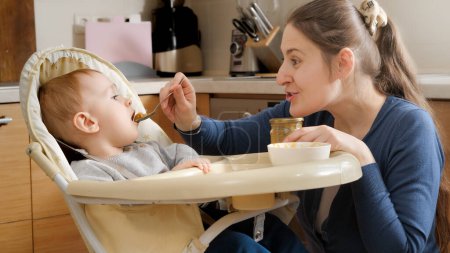 Foto de Portrait of mother playing with her baby son in airplanes while feeding him in highchair. Concept of parenting, healthy nutrition and baby care - Imagen libre de derechos