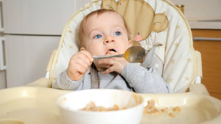 Téléchargez les photos : Portrait de bébé garçon mignon avec les yeux bleus manger de la nourriture avec les mains et la cuillère et obtenir salissant. - en image libre de droit
