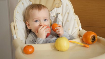 Photo for Portrait of little baby boy sitting in highchair and eating fruits at kitchen. Concept of parenting, healthy nutrition and baby feeding - Royalty Free Image