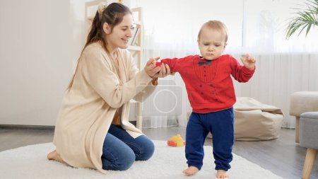 Foto de Una joven sonriente apoya y sostiene a su hijo bebé aprendiendo a caminar sobre una alfombra en la sala de estar. Desarrollo del bebé, juegos familiares, primeros pasos, paternidad y cuidado. - Imagen libre de derechos