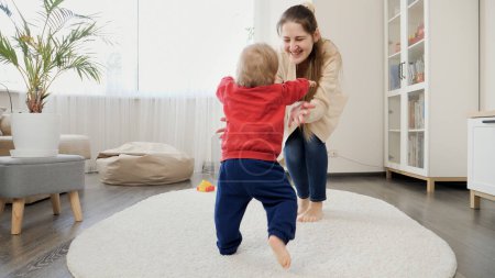 Photo for 1 year old baby boy learning walking and making steps on carpet towards laughing mother. Baby development, family playing games, making first steps, parenthood and care. - Royalty Free Image