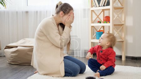 Téléchargez les photos : Joli bébé garçon souriant jouant à cache-cache avec sa mère assise sur le tapis. Développement de bébé, jeux de famille, s'amuser ensemble, parentalité et soins. - en image libre de droit