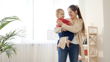 Photo for Beautiful young mother holding her baby boy and showing him toys in living room. Baby development, family playing games, making first steps, parenthood and care. - Royalty Free Image