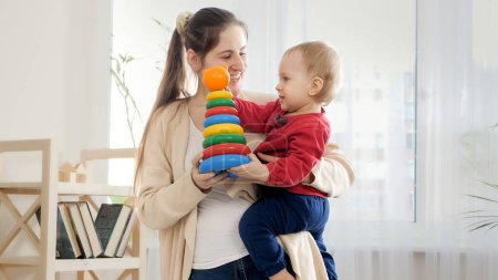 Photo for SMiling mother holding her baby som and giving him colorful toy tower or piramid. - Royalty Free Image