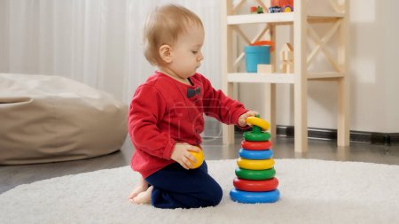 Foto de Pequeño niño de 1 año sentado en el suelo y construyendo una torre de juguete colorida. Desarrollo del bebé, juegos familiares, primeros pasos, paternidad y cuidado. - Imagen libre de derechos