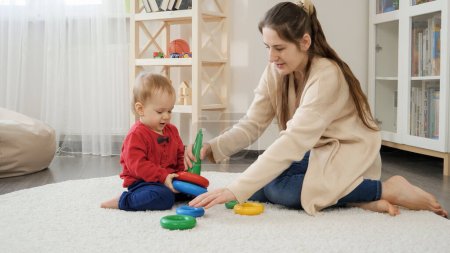 Photo for Beautiful young mother playing with little baby son on floor and assembling toy tower. Baby development, child playing games, education and learning - Royalty Free Image