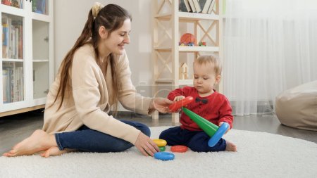 Photo for Young woman helping her baby son building toy tower with colorful rings. Baby development, child playing games, education and learning - Royalty Free Image