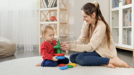 Photo for Young loving mother helping her baby son assembling colorful toy tower or pyramid. - Royalty Free Image