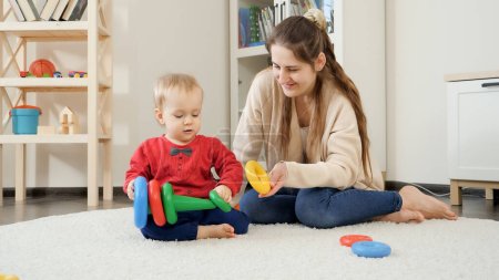 Photo for Young mother helping her baby son and playing with him on carpet in lviing room. - Royalty Free Image