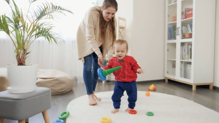 Téléchargez les photos : Joyeux bébé garçon souriant tenant des jouets et apprenant à marcher sur le tapis dans le salon. Développement du bébé, jeux de famille, premiers pas, parentalité et soins. - en image libre de droit