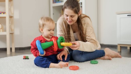 Foto de Joven madre enseñando a su pequeño hijo a construir colorida pirámide de juguetes en casa. Desarrollo del bebé, juegos infantiles, educación y aprendizaje - Imagen libre de derechos