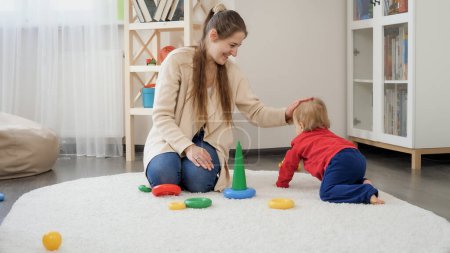 Photo for Happy smiling mother and cute baby boy crawling on soft carpet in living room. - Royalty Free Image