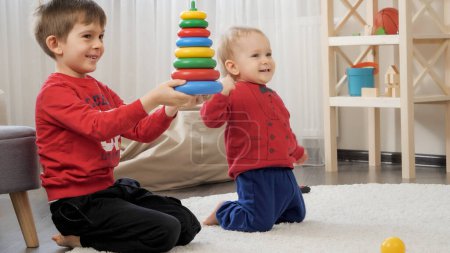 Téléchargez les photos : Deux petits garçons heureux souriant et acclamant après avoir assemblé la tour de jouet sur le tapis dans le salon. Concept d'éducation des enfants, développement du bébé, jeux en famille - en image libre de droit