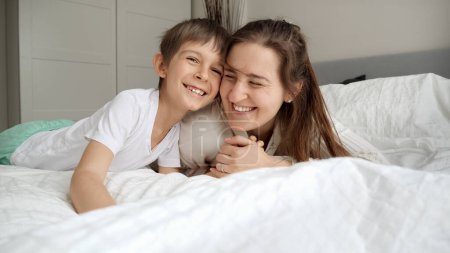 Téléchargez les photos : Joyeux garçon joyeux avec la mère portant un pyjama couché dans le lit et passer du bon temps ensemble. - en image libre de droit