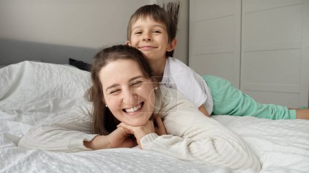 Téléchargez les photos : Portrait d'un petit garçon aimant embrassant sa mère couchée au lit. Concept de famille heureuse, parents avec enfants, émotions positives et détente à la maison - en image libre de droit