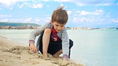 Téléchargez les photos : Garçon assis sur la plage de la mer, creuser et jouer avec le sable. Concept de tourisme, voyage, vacances d'été et week-ends. - en image libre de droit