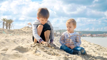 Foto de Dos chicos sentados en la playa de arena y jugando con coches de juguete. Concepto de turismo, viajes, vacaciones de verano. - Imagen libre de derechos