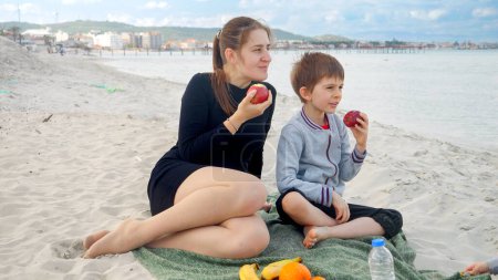 Foto de En un día soleado en la playa, una mujer feliz y su hijo comparten un picnic, comiendo manzanas y relajándose junto al mar. Familia pasar un buen rato juntos al aire libre. - Imagen libre de derechos