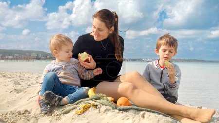 Foto de Retrato de familia feliz con dos niños haciendo picnic en la playa de arena y comiendo frutas. Vacaciones familiares y fin de semana en la naturaleza - Imagen libre de derechos