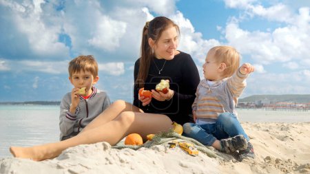Photo for A happy woman enjoys a beach picnic with her two sons, eating fresh fruits and feeling joy. Family vacation and weekend on nature - Royalty Free Image