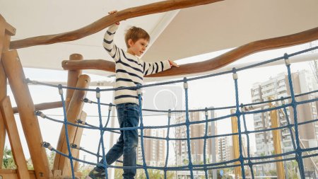 Foto de Niño pequeño balanceándose en el puente de cuerda de madera en el patio al aire libre en el parque. - Imagen libre de derechos