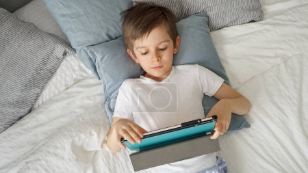 Téléchargez les photos : Heureux jeune garçon joue avec une tablette pendant qu'il est couché dans son lit pendant la journée. Enfant utilisant gadget, éducation et développement - en image libre de droit
