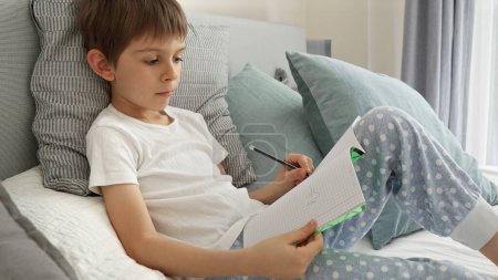 Téléchargez les photos : Portrai de petit garçon travaillant sur ses devoirs au lit, écrivant dans un carnet avec concentration. Importance de l'éducation et du développement de l'enfant dans un environnement d'apprentissage à distance - en image libre de droit