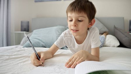 Téléchargez les photos : Jeune garçon en pyjama écrivant dans un cahier alors qu'il était allongé au lit, faisant ses devoirs. Éducation et développement de l'enfant dans un environnement d'apprentissage à distance - en image libre de droit