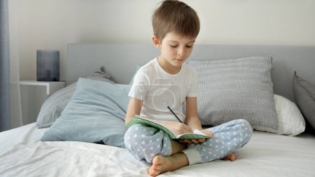 Téléchargez les photos : Un garçon souriant en pyjama faisant ses devoirs au lit le matin. Concept d'éducation, de développement de l'enfant et d'enseignement à distance à domicile - en image libre de droit