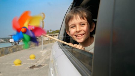 Photo for Smiling boy looking through open car window and holding colorful spinning pinwheel because of blowing wind. Concept of children travel, journey, tourism, holiday, weekend and vacation - Royalty Free Image