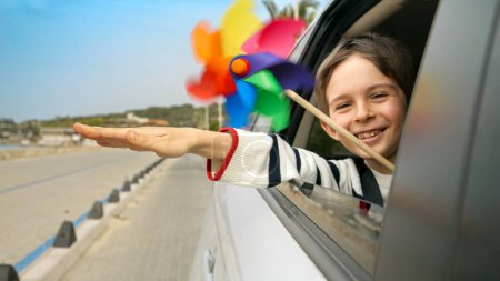 Foto de Niño feliz sosteniendo una paleta de colores o molino de viento mientras mira por la ventana de un coche. Concepto de viaje, viaje, turismo, vacaciones, fin de semana y vacaciones para niños - Imagen libre de derechos