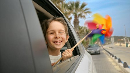 Foto de Feliz niño sonriente sosteniendo paletas de colores o molinete mientras mira por la ventana abierta del coche. Concepto de viaje, viaje, viaje, vacaciones de verano y vacaciones. - Imagen libre de derechos