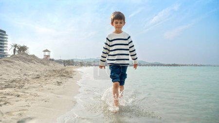 Téléchargez les photos : Joyeux garçon souriant en pull marchant sur la plage de sable fin de la mer et éclaboussant l'eau avec les pieds. Vacances, vacances, week-end à la nature - en image libre de droit