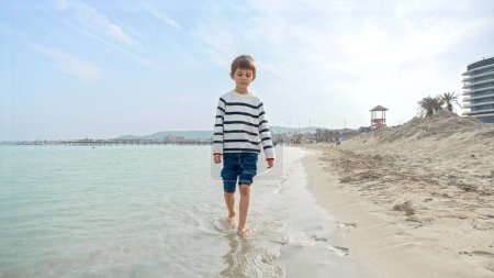 Foto de Lindo niño sonriente en suéter salpicando agua de mar mientras camina en la playa de arena. Vacaciones, vacaciones, fin de semana en la naturaleza - Imagen libre de derechos
