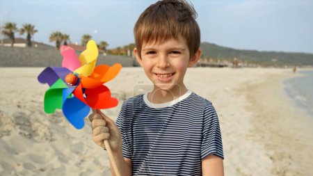 Foto de Lindo chico sosteniendo colorido volante giratorio en la playa de arena del mar. Concepto de vacaciones de verano, felicidad, alegría, viajes y fin de semana - Imagen libre de derechos