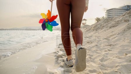 Téléchargez les photos : Mouvement lent de la jeune femme tenant le volant coloré marchant sur la plage de sable fin vers le coucher du soleil. Vacances d'été, voyages et émotions positives - en image libre de droit