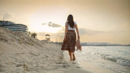 Foto de Mujer joven con bufanda de seda caminando en la playa de arena de mar en el día ventoso hacia la puesta del sol. Concepto de felicidad, viajes, viajes, viajes, turismo y vacaciones - Imagen libre de derechos